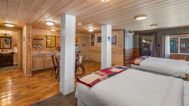 A cabin-style room with wooden floors, two beds, a kitchenette, and a bathroom.
