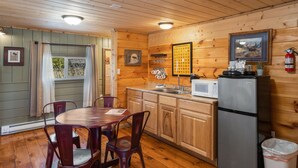 A wooden cabin interior with a dining table, chairs, a kitchen area with a refrigerator, microwave, and a framed U.S. National Parks sign.