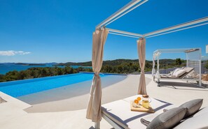 Ein privater Pool mit freiem Blick auf das Meer und die Hügel, ein überdachter Sitzbereich mit Kissen und ein Tisch mit Getränk und Obst.
