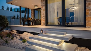 A modern house with a stone exterior, a dining area with outdoor seating, and a staircase leading to the entrance. - Luxury Villa Siesta with Pool (Jezera)