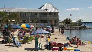 A beach with umbrellas, lounge chairs, and a building with a metal roof. - ⭐️Gorgeous Sunset Island Private Beach In/Outdoor Pools⭐️ (Ocean City)