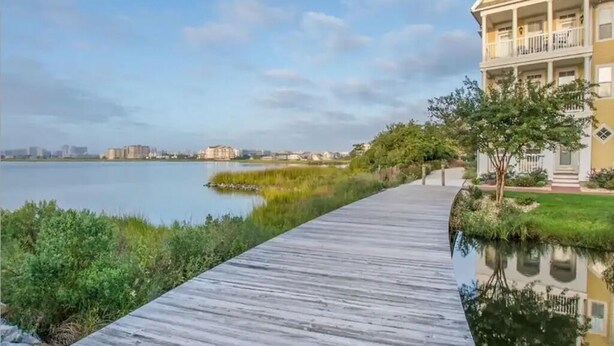 A wooden boardwalk leading to a yellow house with a balcony, overlooking a calm body of water and a distant city skyline. - ⭐️Gorgeous Sunset Island Private Beach In/Outdoor Pools⭐️ (Ocean City)