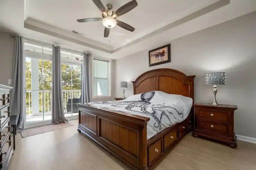 A bedroom with a large bed, a ceiling fan, a nightstand, and a view of the outdoors.