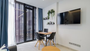 A modern living room with a sofa, a dining table with chairs, a TV mounted on the wall, and a large window with curtains.