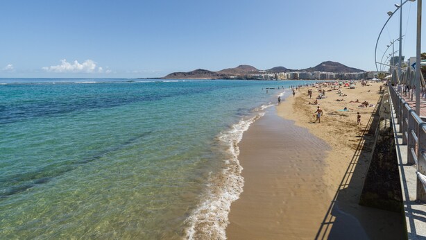 A beach with clear waters, a sandy shore, and a promenade with umbrellas.