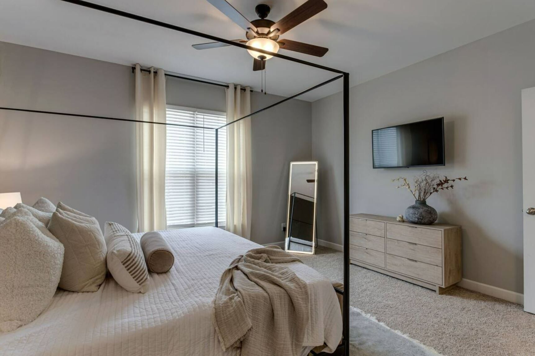 Ein Schlafzimmer mit Bett, Deckenventilator, Kommode, Fernseher und Fenster mit Jalousien.
