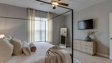 Ein Schlafzimmer mit Bett, Deckenventilator, Kommode, Fernseher und Fenster mit Jalousien.
