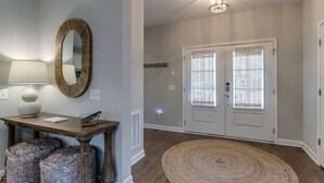 Un couloir avec une console en bois, un miroir, des poufs à motifs, un tapis tissé et une porte à deux vantaux avec des stores.