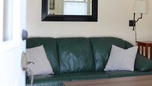 A green leather sofa with striped pillows, a wooden coffee table, a wall-mounted mirror, and a shelf with various bottles and a clock. - Guest House 10 Min to Smugglers! (Cambridge)