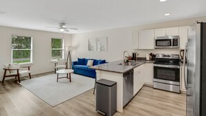 A modern kitchen with white cabinets, a stainless steel refrigerator, a microwave, and an oven. There is a blue couch in the living area and a small island with a sink. - Entire Home: Coastal 3BR Escape with Garage & Pets (St. Augustine)