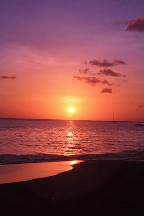 A sunset over a beach with calm waters and a sailboat in the distance. - CHOLSON CHALETS LIMITED (Charlotteville)