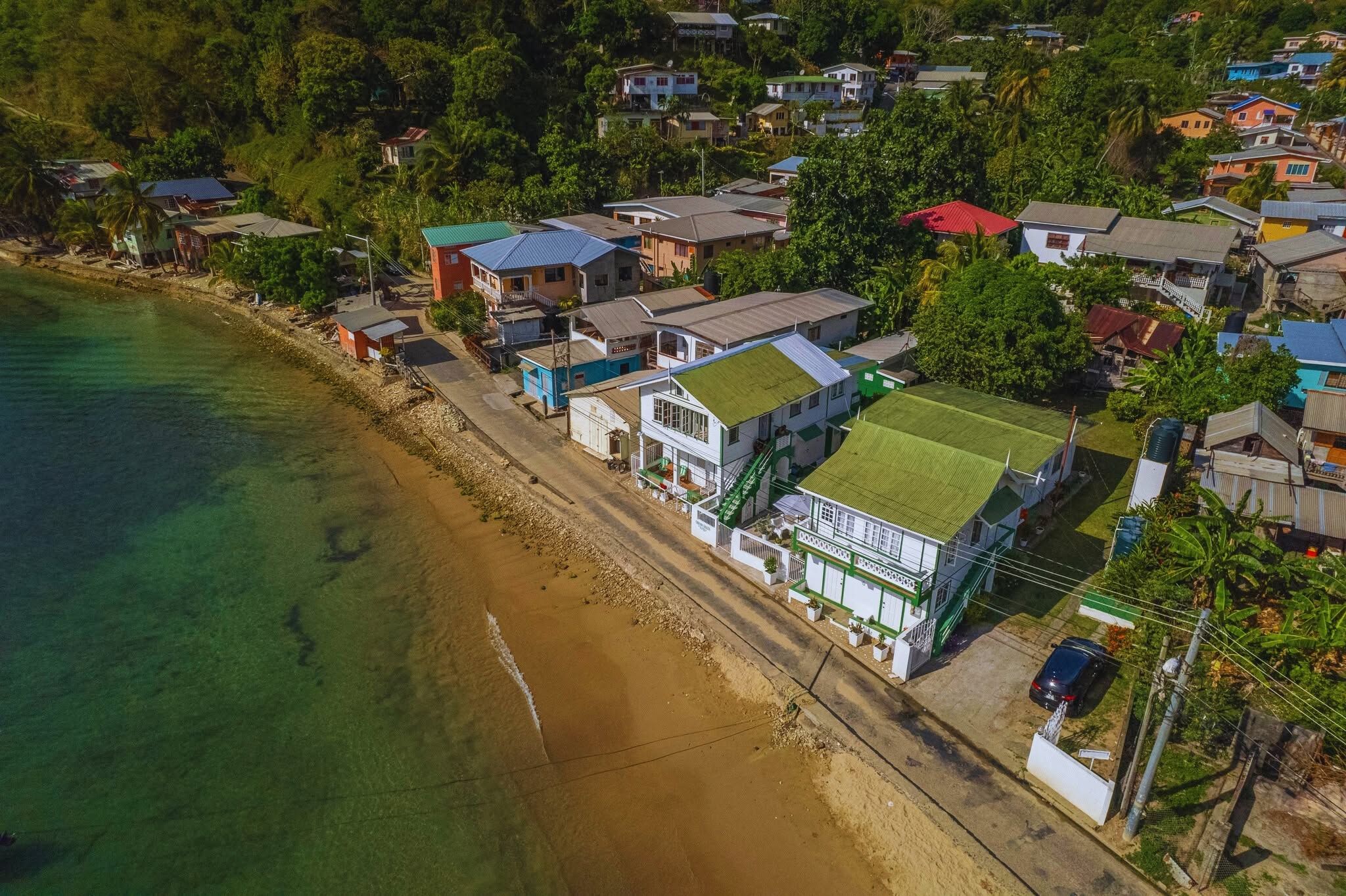 An aerial view of a coastal residential area with houses along the beach, clear water, and dense greenery.