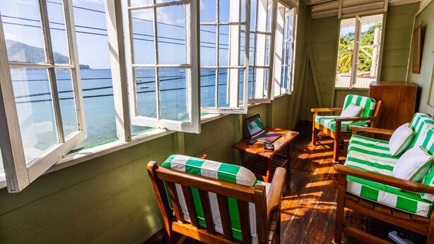 A room with large windows overlooking a body of water, wooden furniture with green and white striped cushions, and a laptop on a small table. - CHOLSON CHALETS LIMITED (Charlotteville)