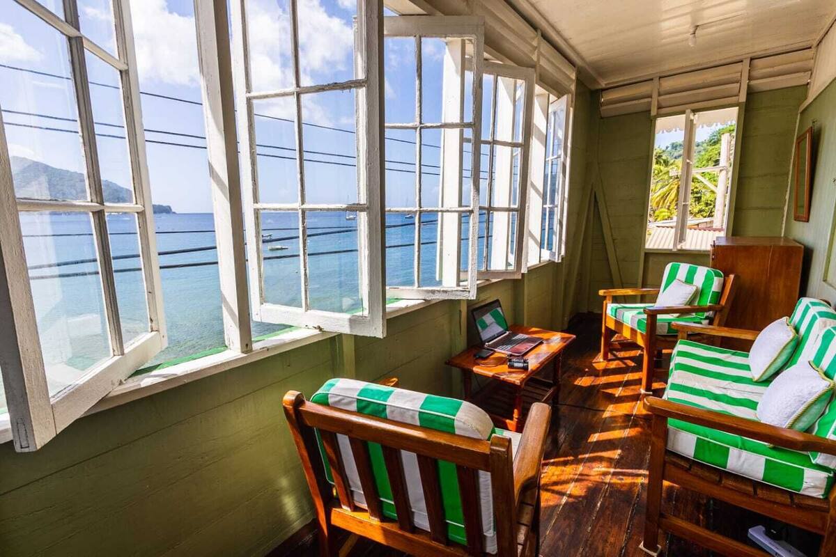A room with large windows overlooking a body of water, wooden furniture with green and white striped cushions, and a laptop on a small table.