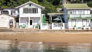 A coastal building with a white facade and green accents, situated by a sandy beach with a backdrop of lush greenery. - CHOLSON CHALETS LIMITED (Charlotteville)