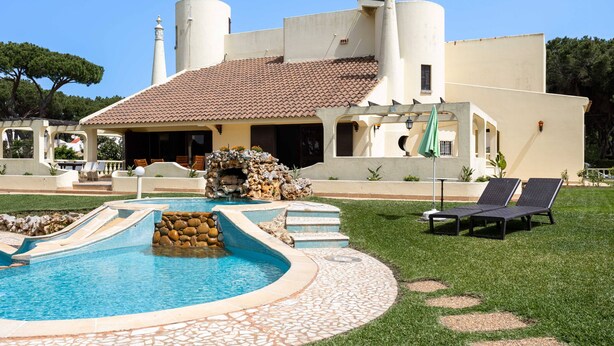 Ein Pool mit gefliestem Rand, einer steinernen Wasserfallanlage und Liegestühlen.