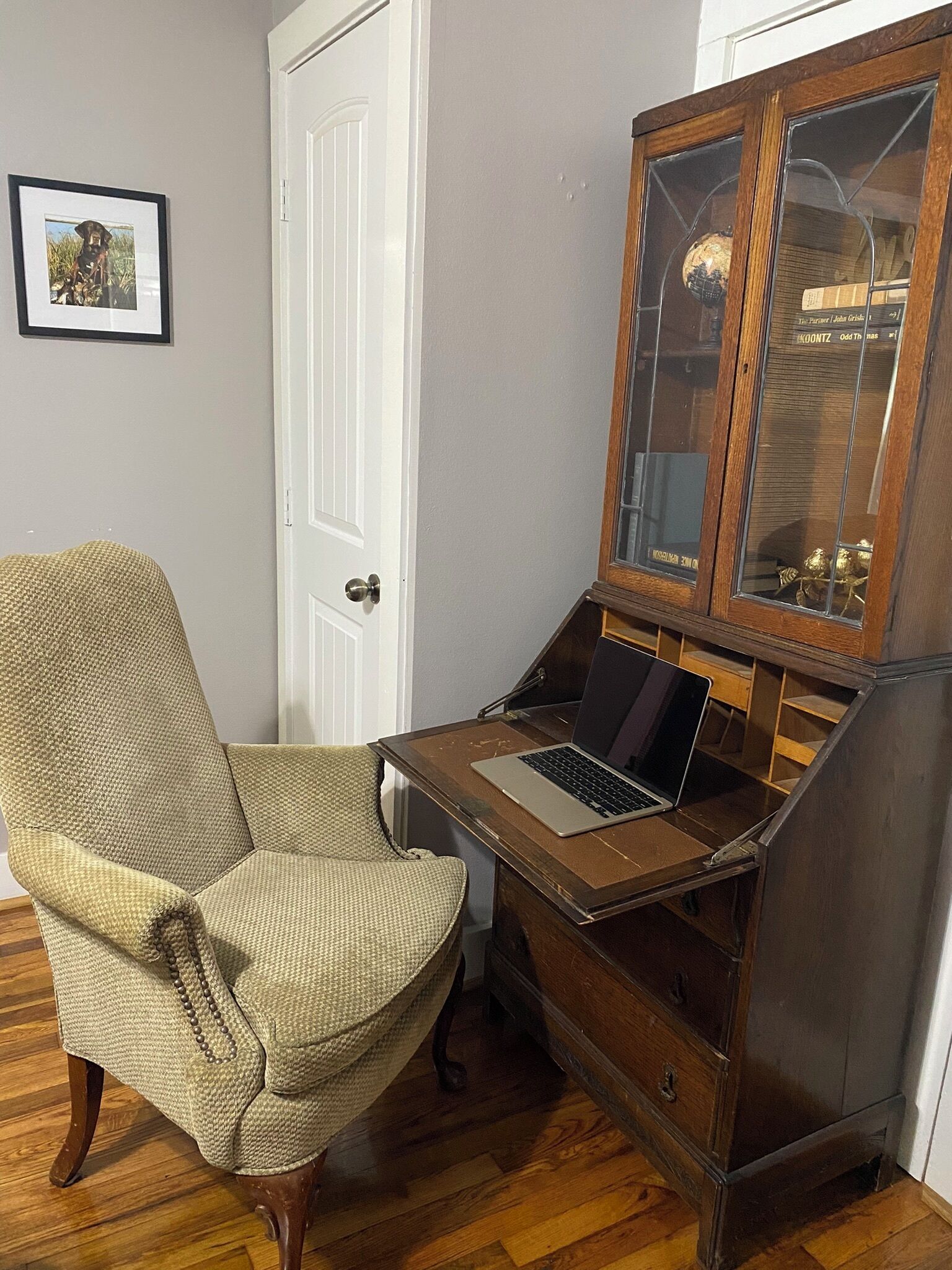 Una habitación con un escritorio de madera, una computadora portátil, una silla de mimbre, un mueble de madera con puertas de vidrio y un cuadro en la pared.