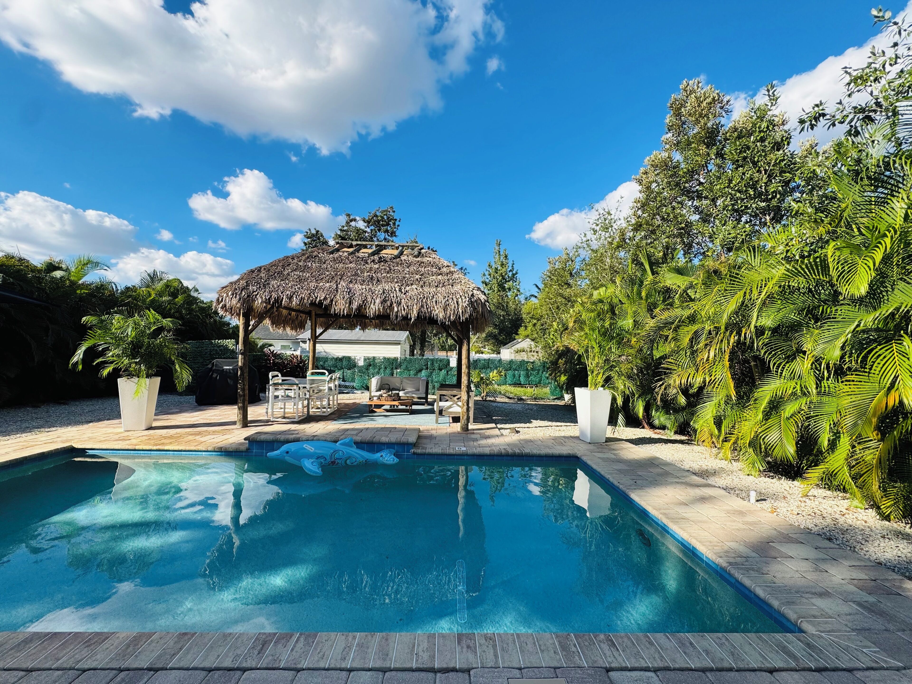 A swimming pool with a thatched gazebo, surrounded by lush greenery and a clear blue sky.