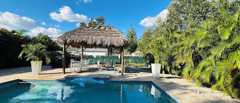 A swimming pool with a thatched gazebo, surrounded by lush greenery and a clear blue sky.