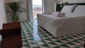 Une chambre d’hôtel avec un lit, une plante en pot, un sol à carreaux verts et blancs, et une vue sur l’extérieur.