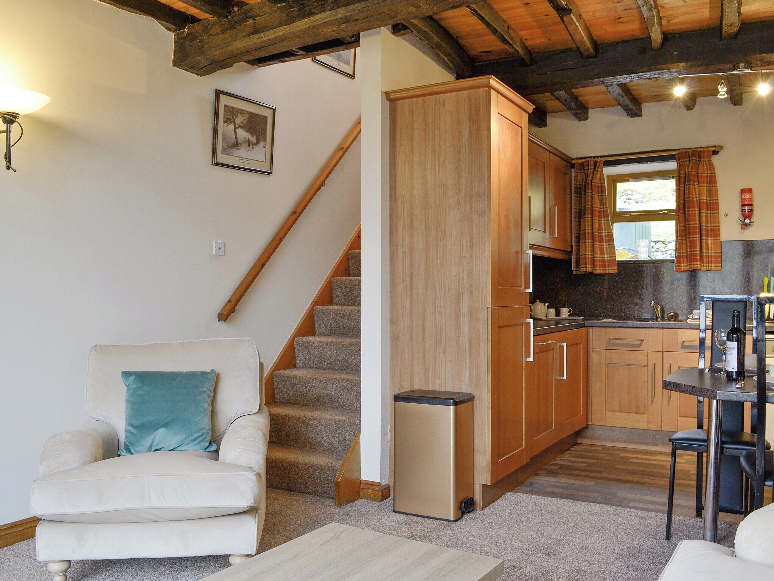 A cozy living room with a sofa, a staircase, and a kitchen area with wooden cabinets and a window with plaid curtains.