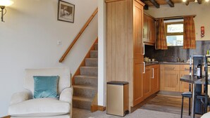 Un salon chaleureux avec un canapé, un escalier et un coin cuisine doté de meubles en bois et d’une fenêtre ornée de rideaux à carreaux.