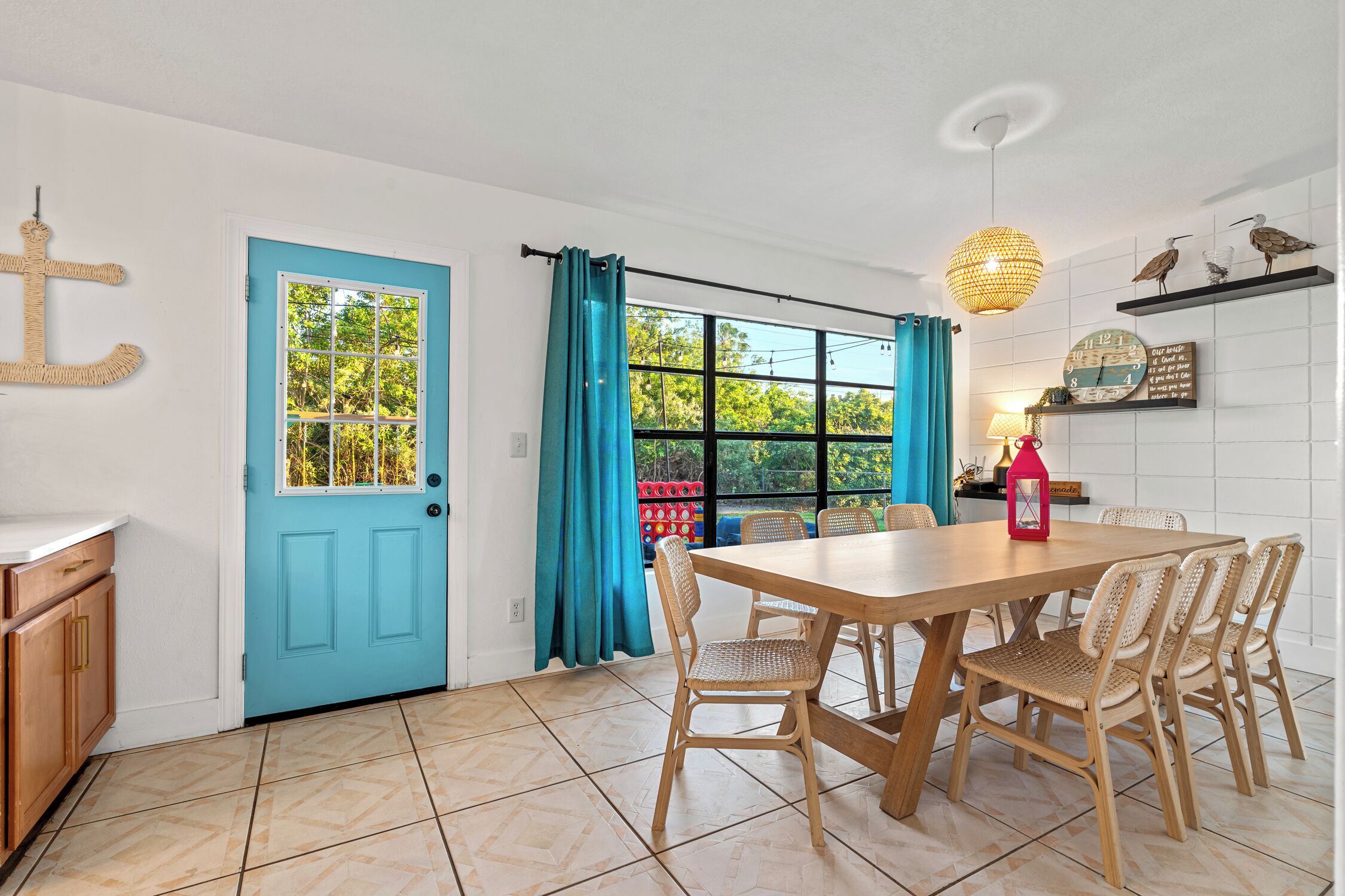 Un coin repas avec une table en bois et des chaises en osier, une porte bleue et une fenêtre avec des rideaux.
