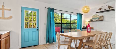 Un coin repas avec une table en bois et des chaises en osier, une porte bleue et une fenêtre avec des rideaux.