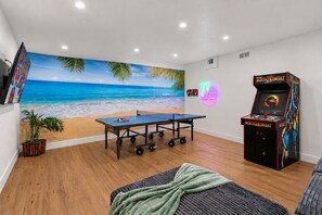 Um quarto com mesa de pingue-pongue, um jogo de arcade Mortal Kombat, um mural de praia, uma placa de palmeira neon e um vaso de planta.