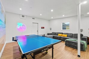Uma mesa de ping pong em um quarto com um sofá e um quadro de uma praia.