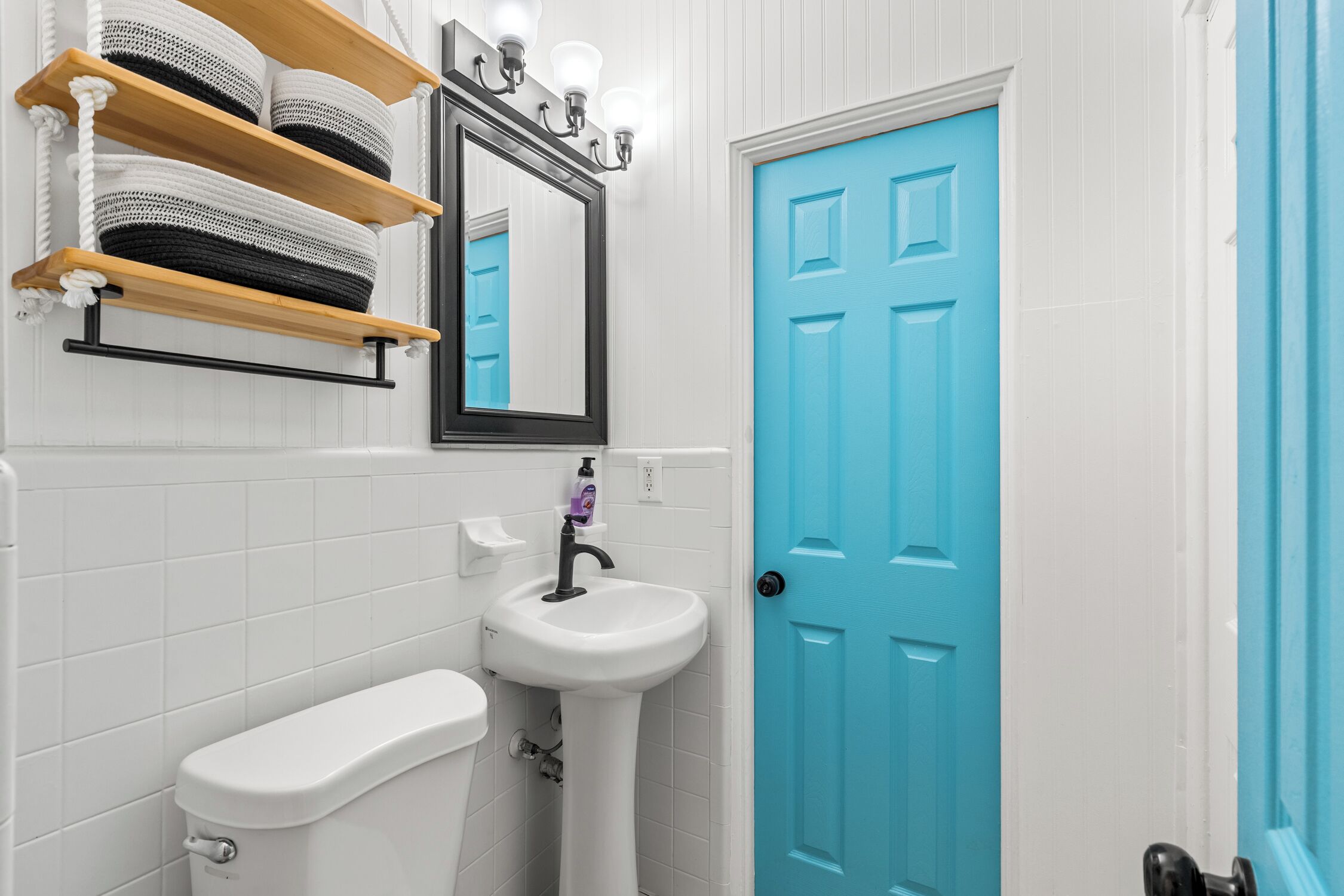 Une salle de bain avec des toilettes blanches, un lavabo blanc, une porte bleue et des étagères avec des paniers.