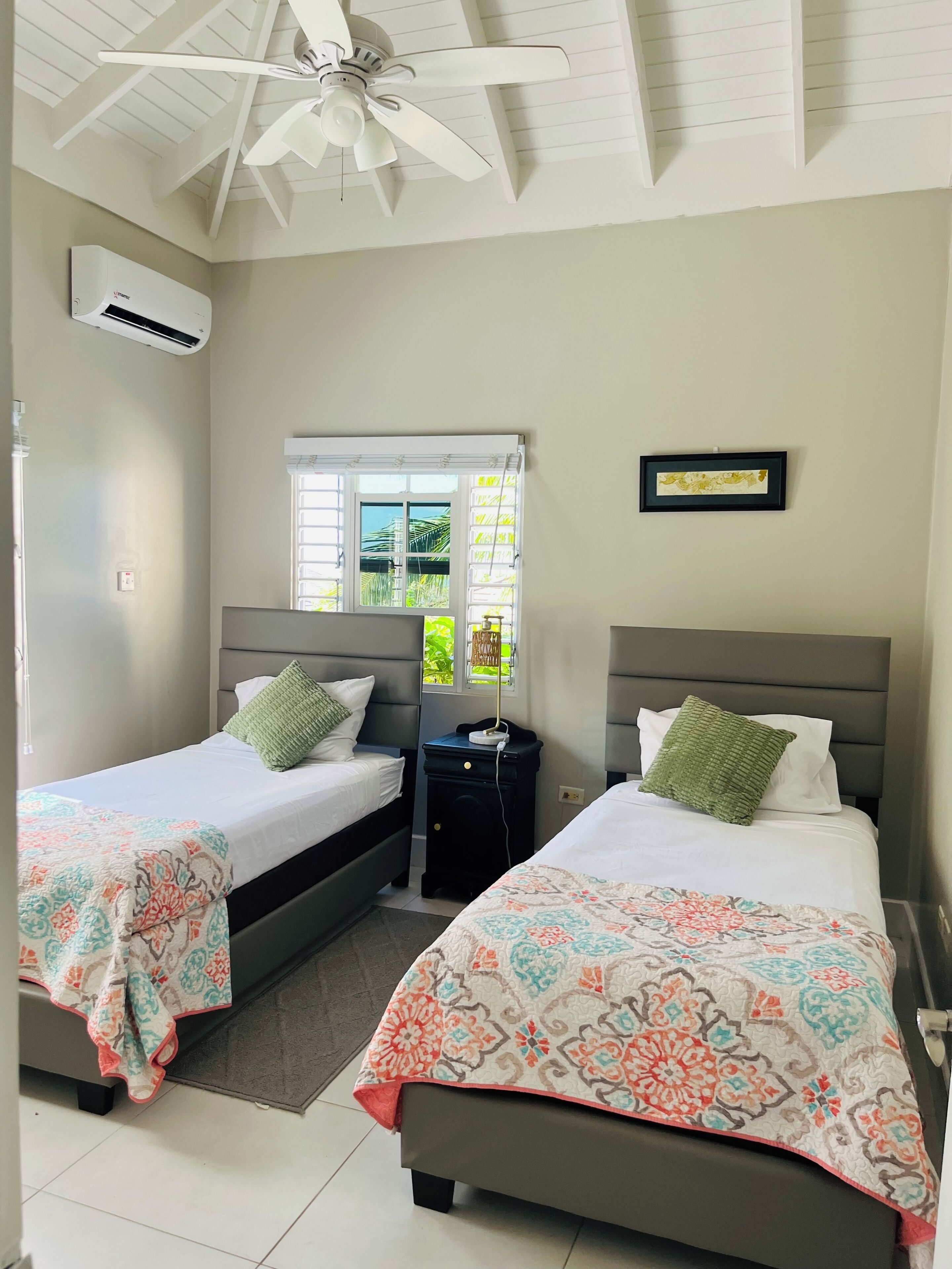 Une chambre d’hôtel avec deux lits, un ventilateur de plafond, un appareil de climatisation, une fenêtre avec des stores, une table de chevet avec une lampe et un tableau encadré au mur.