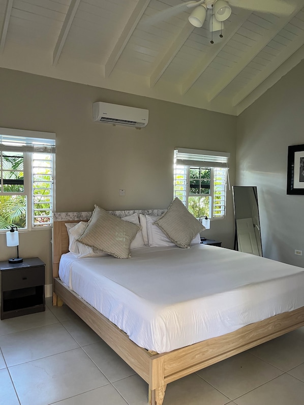 A bedroom with a large bed, two windows with blinds, an air conditioning unit, a bedside table with a lamp, and a mirror on the wall.