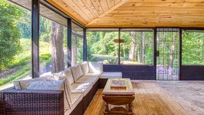 A modern outdoor seating area with a sofa and a wooden table, large windows offering a view of a forested area.