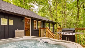 A cabin with a hot tub, a grill, and a wooden deck surrounded by trees.