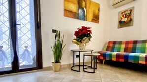 Una habitación con un ventanal grande, un sofá con una manta colorida, una mesita con un jarrón de flores rojas, una planta en maceta y un cuadro en la pared.