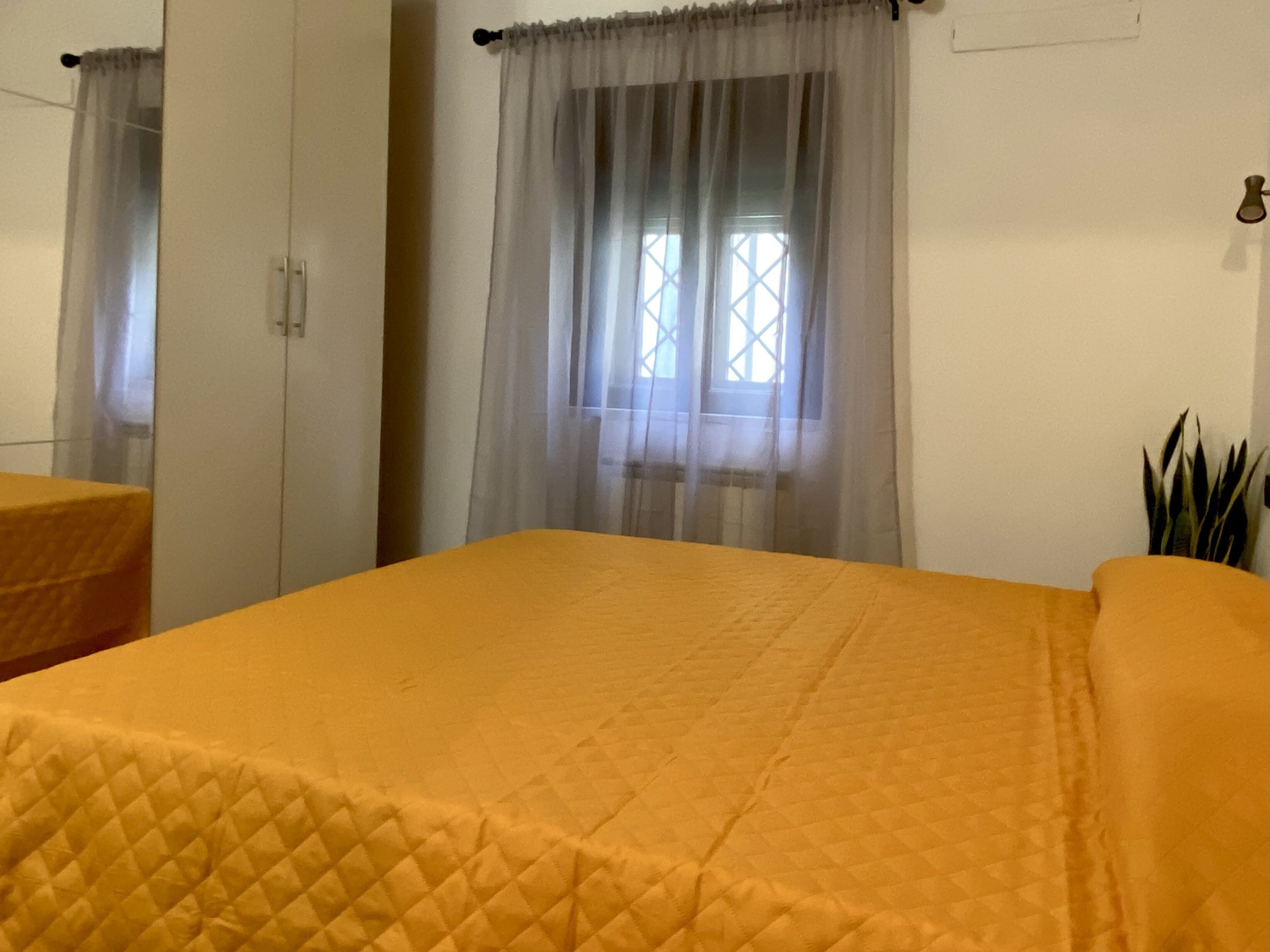 Une pièce avec un lit recouvert d’une couverture matelassée, une fenêtre ornée de rideaux légers et une plante placée sur le côté droit.
