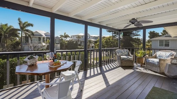 Um terraço coberto ao ar livre com mesa e cadeiras, ventoinha no teto e vista para as casas vizinhas.