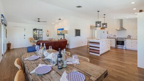 Uma sala de estar ampla com uma mesa de jantar de madeira posta para uma refeição, um canto de estar aconchegante com um sofá vermelho e uma cozinha moderna com armários brancos e eletrodomésticos de aço inoxidável.