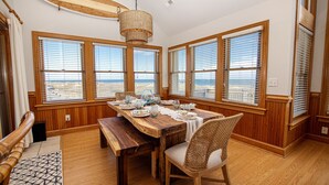 Un coin repas avec une table en bois dressée pour quatre personnes, de grandes fenêtres équipées de stores et une chaise en osier.
