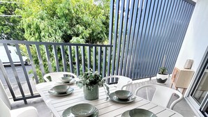 Un balcón con una mesa blanca para cuatro personas, sillas blancas alrededor y una planta en maceta sobre la mesa.