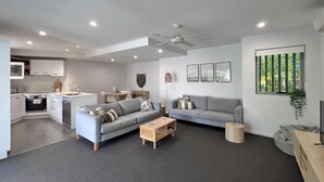 Un espacio de sala moderno con un sofá gris, una mesa de centro de madera y un sofá gris. Se observa una zona de cocina con armarios blancos y una mesa de comedor. Un ventilador de techo es visible, y una ventana ofrece vistas a la vegetación exterior.