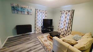 Una sala de estar con una televisión de pantalla plana sobre un mueble bajo, un sofá, una mesa de centro, cortinas estampadas y un cuadro en la pared.