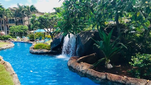 A swimming pool with a waterfall feature, surrounded by lush greenery and palm trees. - Makai Manor Ocean View, sleeps 6. (Kapaʻa)