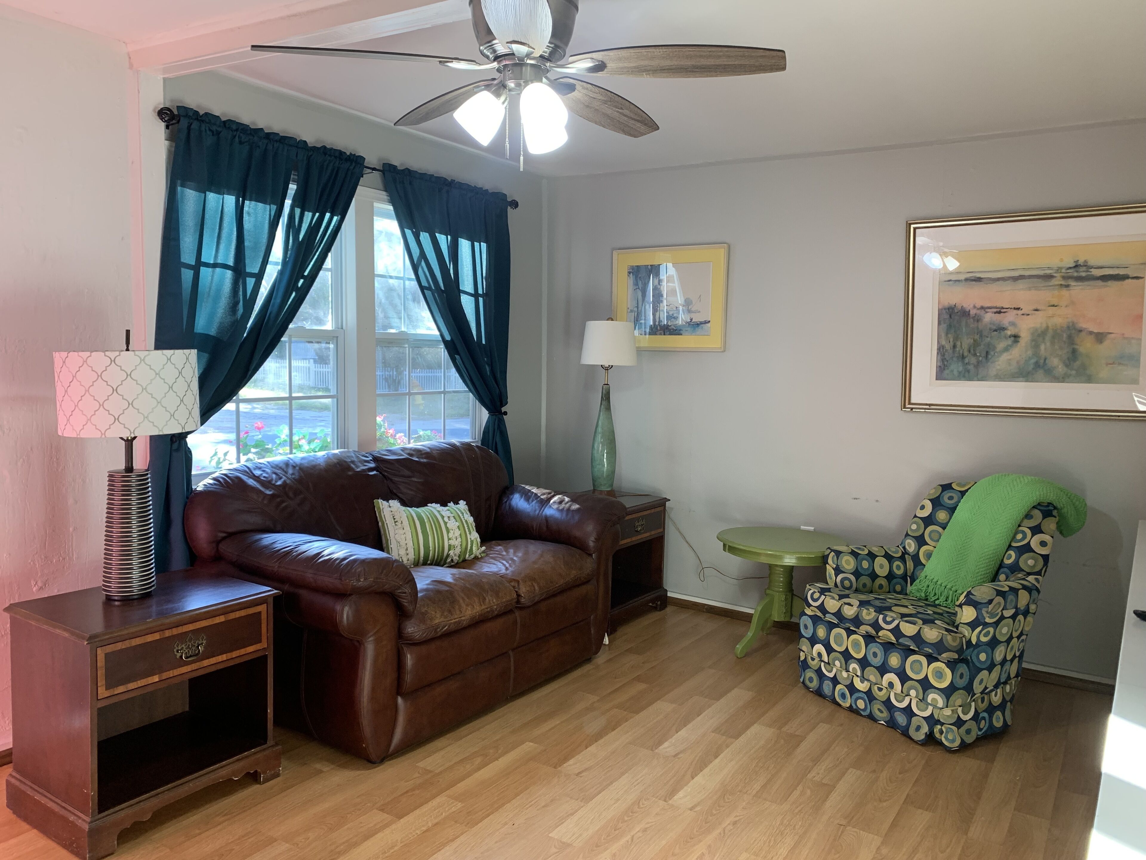 A living room with a brown leather sofa, a green patterned armchair, a wooden side table, a floor lamp, and a ceiling fan.