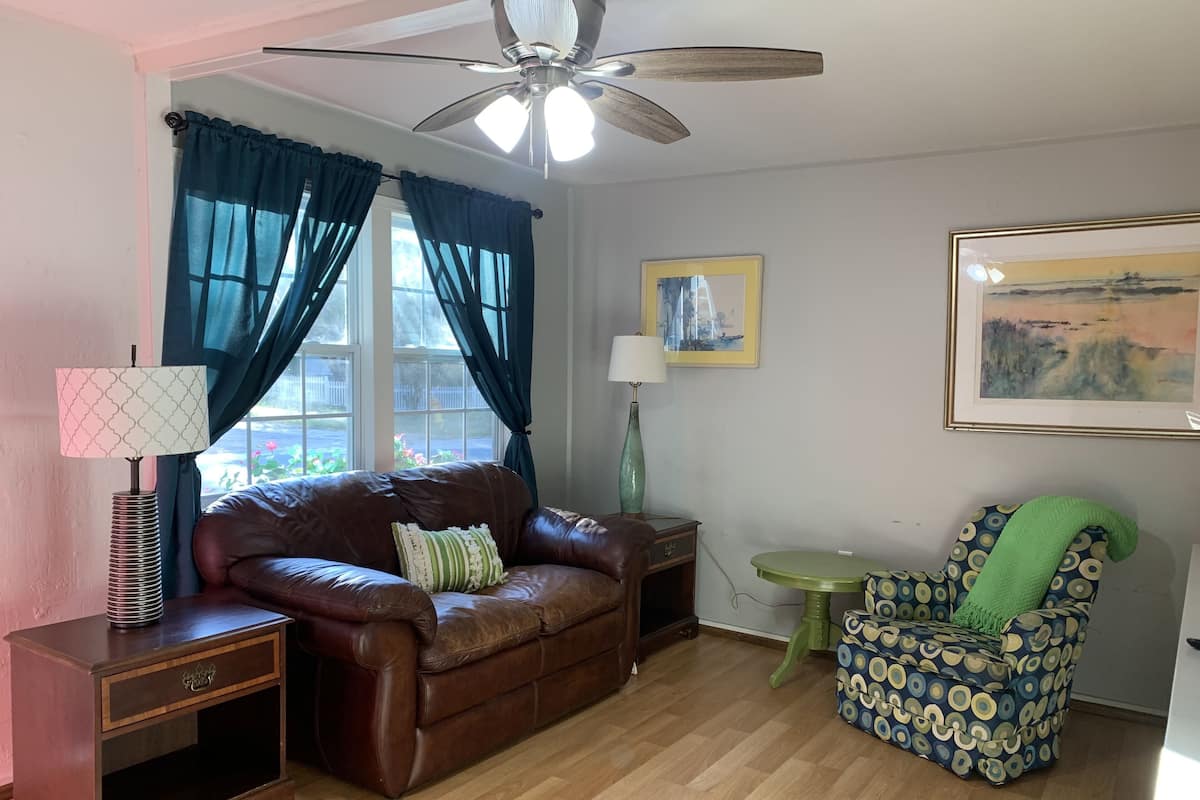 A living room with a brown leather sofa, a green patterned armchair, a wooden side table, a floor lamp, and a ceiling fan.