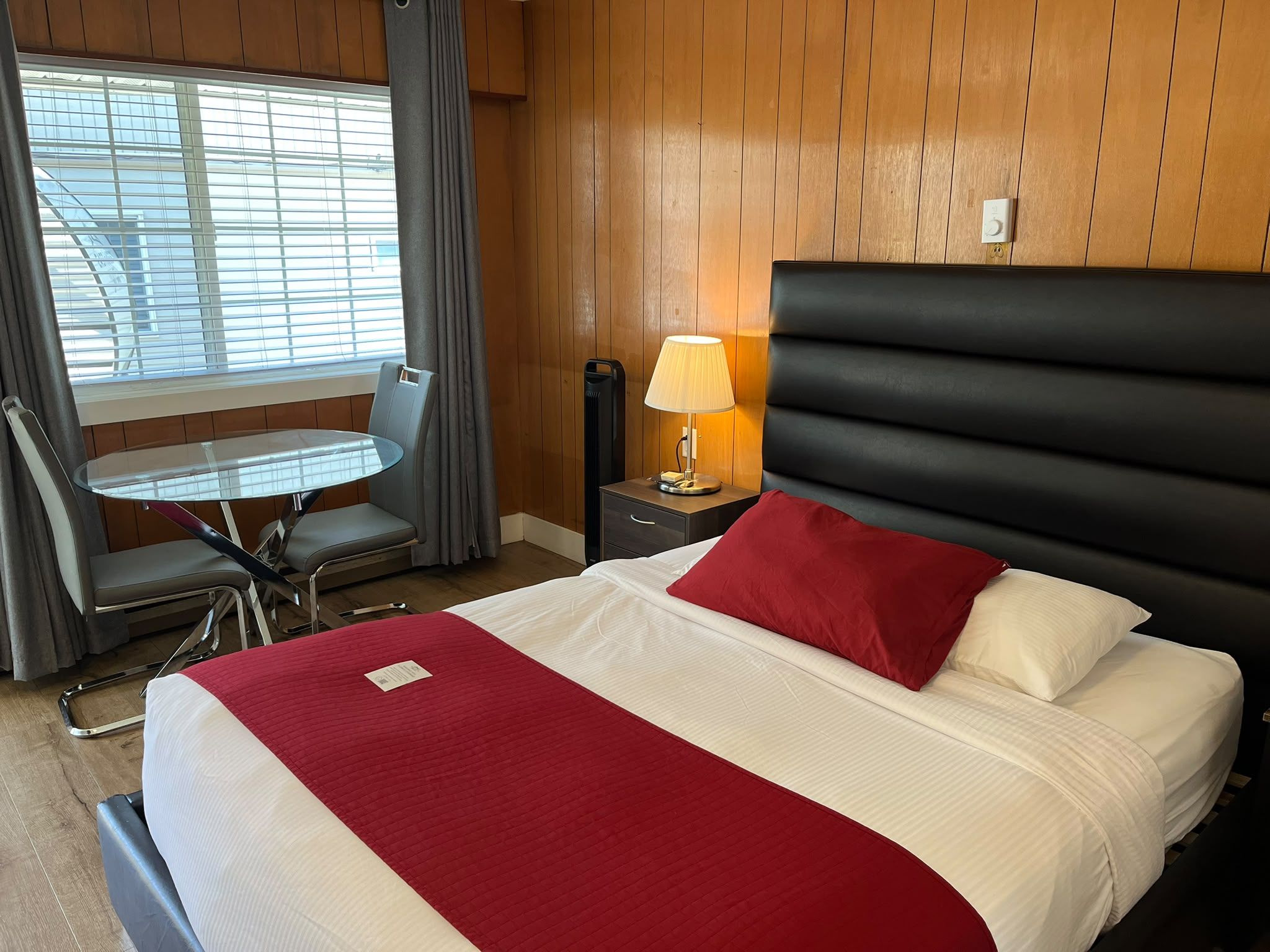 A hotel room with a bed, a bedside table with a lamp, a chair, and a window with blinds.