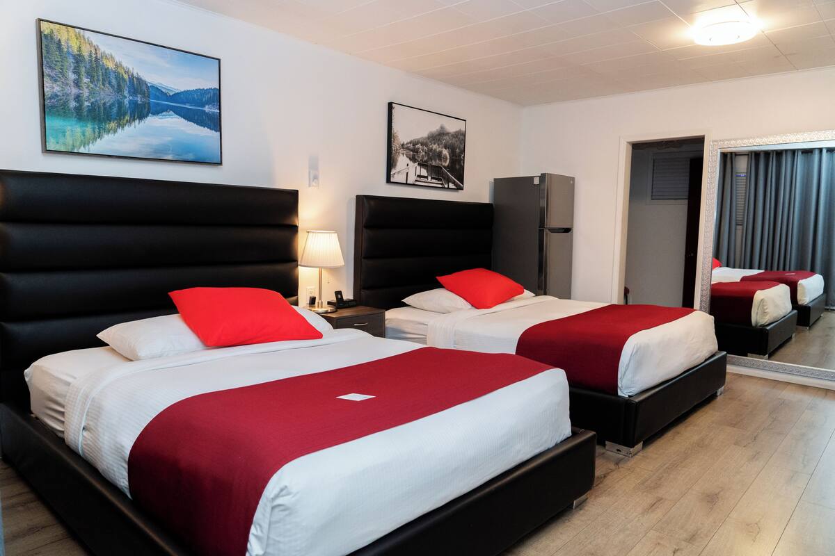 Habitación de hotel con dos camas, cada una con cabecero negro y ropa de cama roja. Se observa un espejo grande en la pared, un televisor de pantalla plana y un cuadro enmarcado sobre las camas.