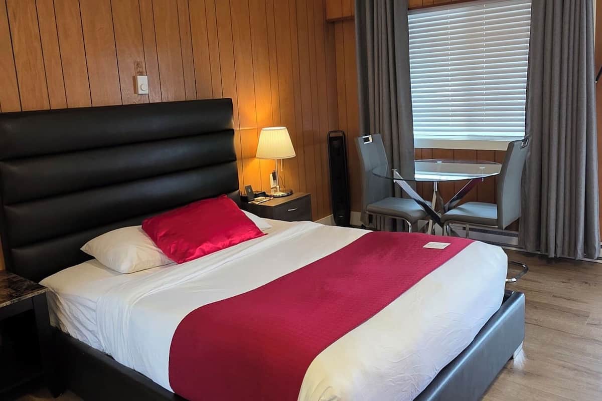 Una habitación de hotel con pared de paneles de madera, una cama tapizada de negro, un escritorio con lámpara y un rincón de comedor con una silla y mesa.