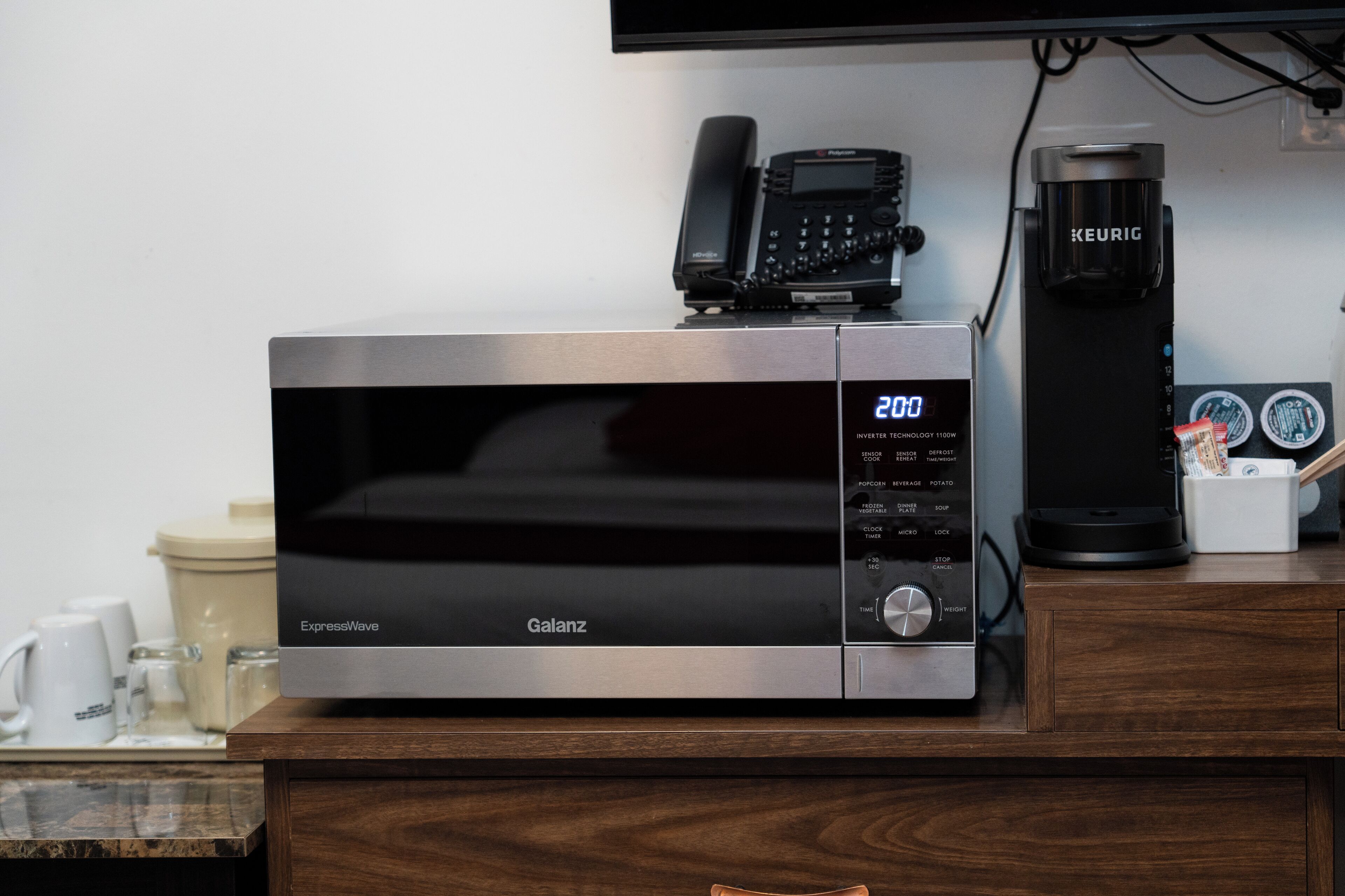 Eine kompakte Mikrowelle mit digitaler Anzeige, eine Kaffeemaschine und ein Telefon auf einer Holztischplatte.